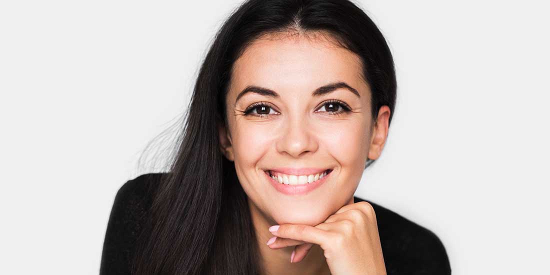 Portrait of brunette cute woman with beautiful and healthy toothy smile, with hand on chin. Young happy pretty woman wearing black sweater on light grey background. Positive and happiness mood.